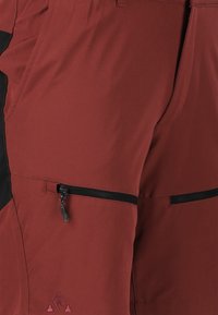Punaiset ja mustat ulkokäyttöön tarkoitetut shortsit, jotka on valmistettu kestävästä kankaasta. Shortseissa on vetoketjullinen tasku ja logo yksityiskohta helmassa.