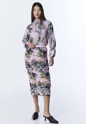 Donna che indossa una camicia a maniche lunghe rosa con motivo e una gonna midi coordinata con texture, abbinata a mules nere con tacco, su sfondo bianco.