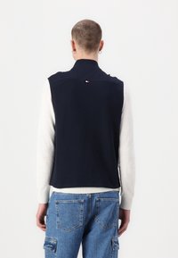 Granatowa pikowana kamizelka z wysokim kołnierzem, zestawiona z jasnoszarym, długim rękawem. Posiada małe logo w trzech kolorach na plecach.