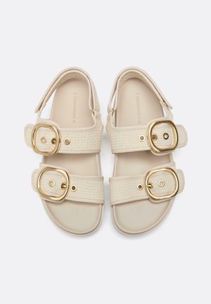 Beige Sandalen mit jeweils zwei breiten, geflochtenen Riemen, großen goldenen Schnallen und gepolsterten Sohlen, nebeneinander auf hellem Hintergrund gelegt.