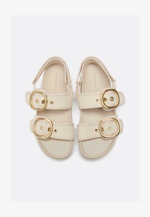 Beige Sandalen mit jeweils zwei breiten, geflochtenen Riemen, großen goldenen Schnallen und gepolsterten Sohlen, nebeneinander auf hellem Hintergrund gelegt.