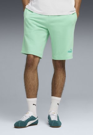 Man met mintgroene Puma-short, witte Puma-sokken en turquoise Puma-sneakers, staand met handen in de zakken.