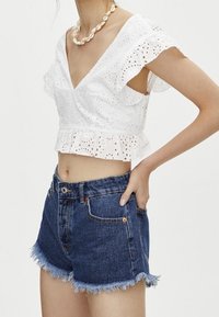 Top corto blanco de eyelet con mangas con volantes y escote en V, combinado con pantalones cortos de mezclilla azules de tiro alto con dobladillo deshilachado y bolsillos.