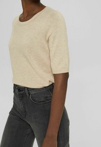 Pull beige à manches courtes avec un col rond, fabriqué en tissu doux et texturé, associé à un jean taille haute gris foncé.