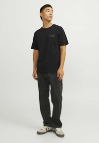 Schwarzes T-Shirt mit einem dezenten "JACK & JONES"-Logo auf der linken Brust, kombiniert mit dunkelgrauen Jeans und hellen Sneakern.