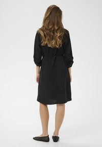 Rochie neagră până la genunchi, cu mâneci trei sferturi și talie înnodată. Textura pare ușoară. Pantofi balerini negri din plasă completează ținuta.