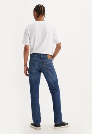 Man met gevlochten haar staat met de rug naar de camera, draagt een wit T-shirt, blauwe spijkerbroek en zwarte sneakers tegen een effen achtergrond.