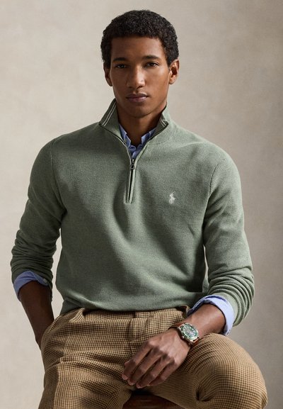 Roheline kudumiga neljandiku tõmblukuga kampsun soonikkoes mansettidega, kantud hele sinise kraega särgi peal; kombineeritud beežide ruuduliste pükstega ja käekellaga.