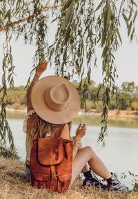 Mochila de cuero marrón con solapa plegable, múltiples bolsillos delanteros y un acabado texturizado. Rodeada de vegetación junto a un cuerpo de agua tranquilo.