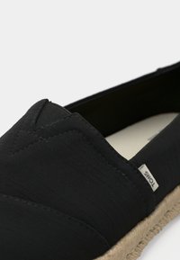 Svart espadrillesko med ovandel i kanvas, texturerad ytfinish och sula av jute rep. Inkluderar en liten varumärkesetikett på sidan.