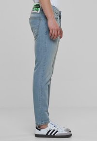 Vaaleansiniset farkut, joissa on kapea malli, viidellä taskulla ja vihreällä tarralla takana. Yhdistettynä valkoisiin lenkkareihin, joissa on mustia yksityiskohtia.