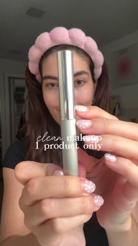 Metallisch silberner Makeup-Applikator, der von einer Person mit hellrosa Akzentnägeln gehalten wird. Weiches rosa Stoff-Haarband, minimale Hintergrunddetails.