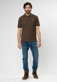 Braunes Polohemd mit kurzen Ärmeln, ausgestattet mit einem Kragen und einem kleinen Logo. Kombiniert mit blauen Jeans und braunen Schnürstiefeln.