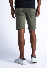 Pantalones cortos verde oliva con dobladillos enrollados y dos bolsillos traseros. Combinados con zapatillas blancas con detalles verdes. Fondo liso.