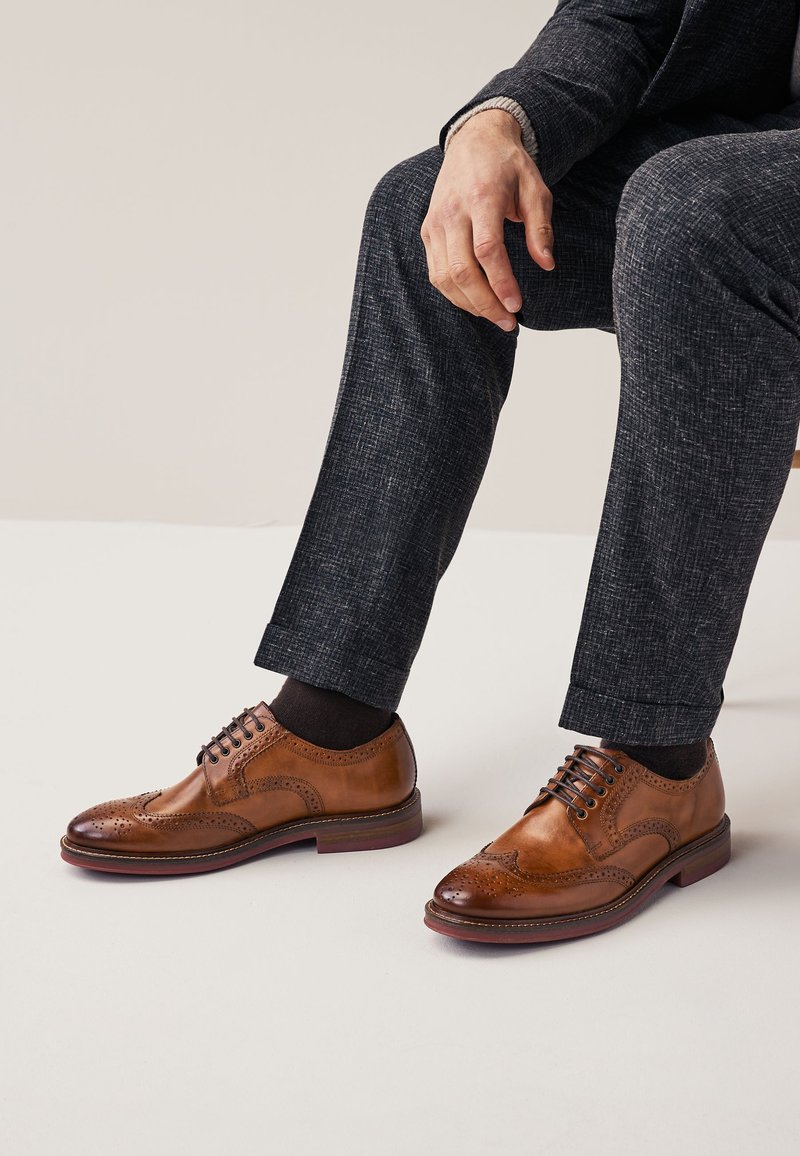 Homme assis, une main reposant sur le genou, portant un pantalon texturé foncé, des chaussettes noires et des chaussures brogues en cuir marron poli.