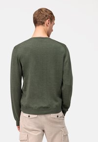 Groene gebreide trui met lange mouwen, ronde hals en geribbelde zoom. Draagtas over beige corduroy broek met zijzakken.