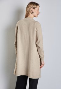 Beige gebreide cardigan met lange mouwen, ribbelrand en manchetten, en zijsplitten. Glad textuur, relaxte pasvorm. G gedragen over zwarte broek.