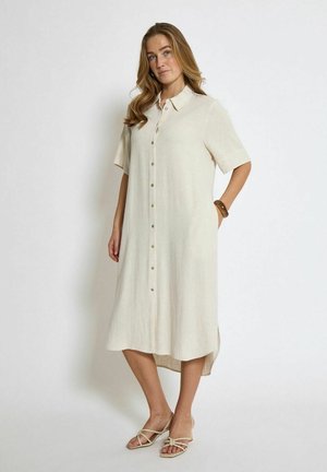 Beige button-up dress made of lightweight fabric, featuring short sleeves, side slits, and a relaxed fit. Worn with neutral sandals.