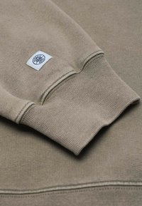 Beige striktrøjeærme med ribbet manchet og lille hvid mærketikette med et cirkulært logo og tekst.