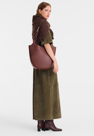 Brown leather shoulder bag with a smooth texture and curved shape, paired with a long olive-green dress and brown ankle boots.