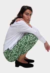 Chemise blanche à manches longues, pantalon à motif floral vert et chaussures à talon carré noires. Modèle en position accroupie, les mains reposant sur les genoux.