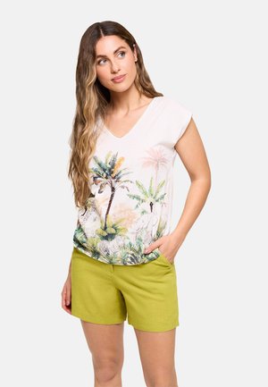 Mujer con cabello largo y ondulado, vistiendo una camiseta blanca con cuello en V y estampado tropical, y pantalones cortos verdes, una mano en el bolsillo, mirando hacia la derecha.
