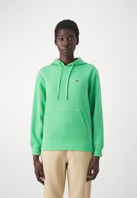 Persona che indossa un felpa verde brillante con tasca frontale e logo Lacoste, abbinata a pantaloni beige, in piedi contro uno sfondo bianco semplice.