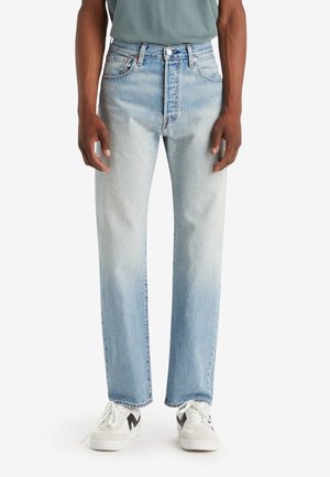 Homme portant un jean droit bleu clair délavé et des baskets blanches avec des accents noirs, debout devant un fond uni.