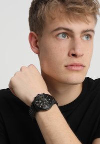 Schwarze Chronographuhr mit Metallarmband, rundem Gehäuse und einem strukturierten Zifferblatt mit silbernen Akzenten und drei Ziffernblättern.