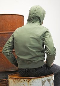 Veste à capuche verte avec une texture lisse légèrement brillante, ourlet et poignets côtelés, portée avec un jean foncé, vue de dos devant un baril rouillé.