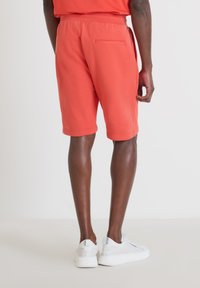 Pantalones cortos rojos hasta la rodilla con una cinturilla elástica y un bolsillo en la parte trasera. Combinados con zapatillas blancas sobre un fondo claro.