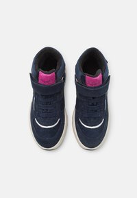 Pár dětských tenisek v námořnické modři s bílou podrážkou, růžovými štítky na jazyku a suchými zipy, zobrazený z výše na bílém pozadí.