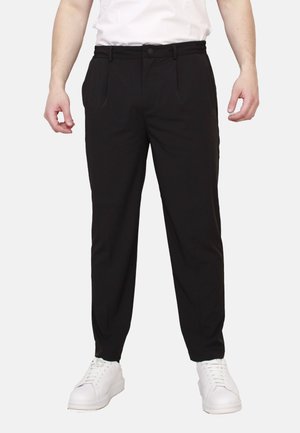 Vista frontale di una persona che indossa pantaloni neri slim fit e sneaker bianche, in piedi davanti a uno sfondo bianco uniforme.