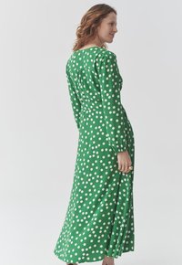 Langes grünes Kleid mit weißen Punkten, ausgestattet mit langen Ärmeln, einem runden Ausschnitt und einem Schlitz am Saum. Glattes Material mit einem leichten Glanz.