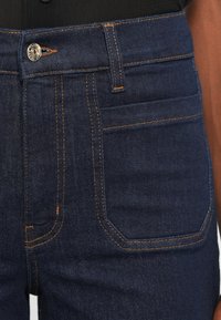 Sötétkék farmernadrág narancssárga cérnával, gombos záródással, övhurkokkal és két hátsó zsebbel. Sima textúra és testhezálló fazon.