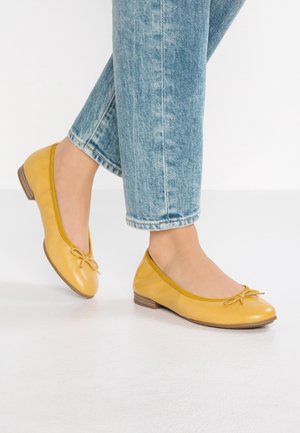 Żółte baleriny ze skóry z zaokrąglonym noskiem, małą kokardką oraz niskim brązowym drewnianym obcasem, zestawione z jasnoniebieskimi jeansami o skróconej długości.