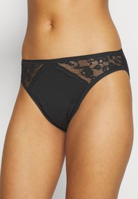 Perizoma nero realizzato in tessuto morbido con accenti di pizzo sui lati, caratterizzato da dettagli a bordo ondulato. Design basic a vita bassa.