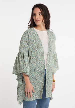 Donna che indossa una giacca stile kimono verde con motivo floreale sopra una blusa bianca traforata e jeans blu, in piedi e guarda di lato.
