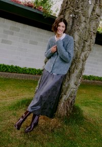 Cardigan grigio a maglia con bottoni, indossato sopra una lunga gonna scura e liscia. Il modello indossa stivali marroni al ginocchio con punta, mentre si appoggia a un albero.
