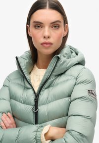 Chaqueta impermeable verde claro con capucha, con cierre de cremallera y puños acanalados, acompañada de un suéter crema debajo.