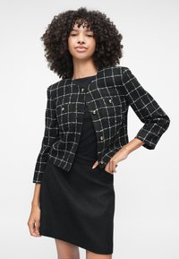 Sort, ternet jakke med hvidt rutemønster og guldknapper, parret med en sort nederdel. Materialet ser tekstureret ud.