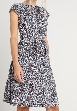 Kobieta w granatowej sukience w kwiaty sięgającej do kolan, z krótkimi rękawami i materiałowym paskiem zawiązanym w talii, stojąca na jednolitym tle.