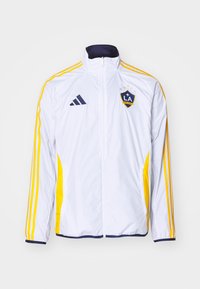 Chaqueta atlética blanca con acentos en azul marino, rayas amarillas y el logo de LA Galaxy. Presenta un cuello alto y bolsillos frontales con cremallera.