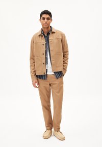 Beige canvas jasje met twee voorzakken over een blauw-wit gestreept shirt. Beige broek en lichtbruine instapschoenen maken de outfit compleet.