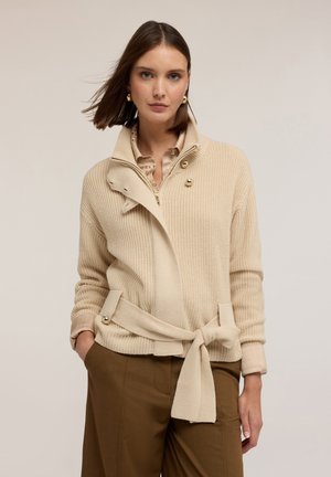 Cardigan beige a costine con colletto ripiegato, bottoni frontali e cintura annodata in vita. Indossato sopra una camicia leggera, abbinato a pantaloni marroni.