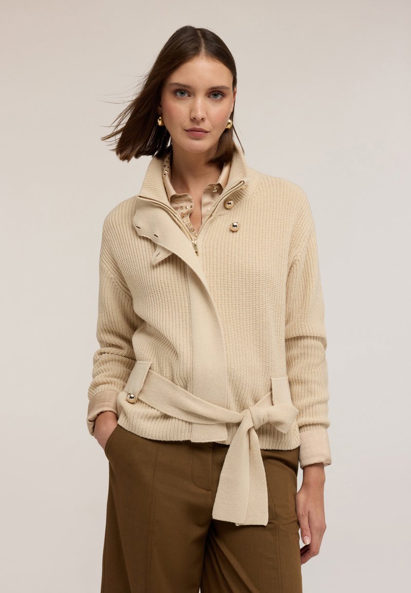 Béžový, žebrovaný pletený cardigan s přeloženým límečkem, předními knoflíky a zavázaným opaskem v pase. Přes lehkou košili, v kombinaci s hnědými kalhotami.