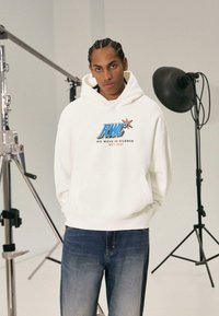 Fehér kapucnis pulóver "RWC" logóval kékként és narancssárgaként, párosítva kék, elhalványult farmernadrággal. Stúdiókörnyezet világítótestekkel és támogató állványokkal.