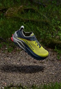 Chaussure de trail running avec un dessus en maille jaune, des accents gris et une épaisse semelle noire. Présente un design rembourré et des lacets colorés.