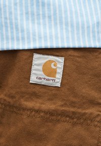 Καφέ ύφασμα με ραμμένη ετικέτα λογότυπου Carhartt, κάτω από ύφασμα με ανοιχτό μπλε και λευκές ρίγες.