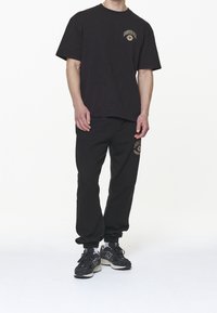 Černá bavlněná sada t-trička a tepláků, s malým vyšitým logem ve zlaté barvě. Volný střih s kulatým výstřihem a elastickými manžetami.